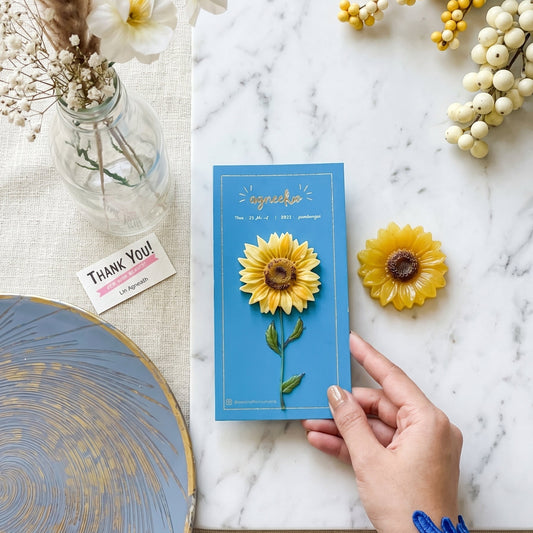 Hand holding a blue card with a sunflower design on a marble surface with flowers and a vase.